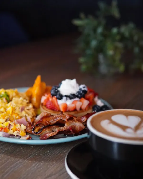 Un plato de brunch con huevos, tocineta y pancakes junto a un latte en O Positivo
